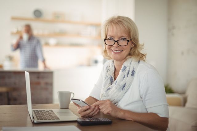 Senior woman sitting at table using mobile phone and laptop, smiling. Ideal for illustrating concepts of senior citizens embracing technology, remote work, modern lifestyle, and digital communication.