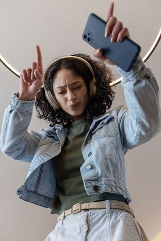 Asian woman dancing and recording with phone and headphones under ring light, denim jacket
