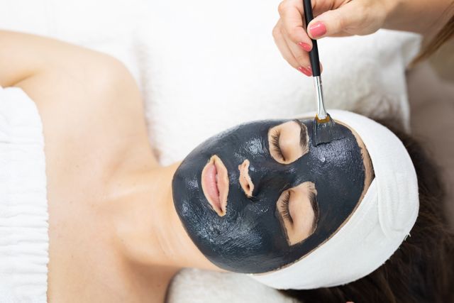 Beautician applying a black face mask to a relaxed female customer at a beauty salon. Ideal for use in articles or advertisements related to skincare, beauty treatments, spa services, and wellness. Perfect for promoting beauty salons, skincare products, and self-care routines.