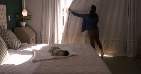 Mother opening curtains to let daylight illuminate sleeping child