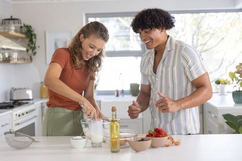 Couple cooking together on sunlit kitchen island whisking batter and laughing