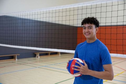 Smiling teenage male holding red white blue volleyball by gym net wearing blue shirt