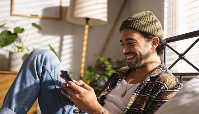 Young man relaxing on bed texting with smartphone wearing beanie and patterned shirt