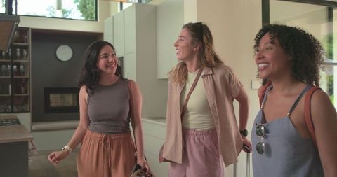 Diverse female friends arriving at bright open-plan kitchen, laughing and carrying luggage