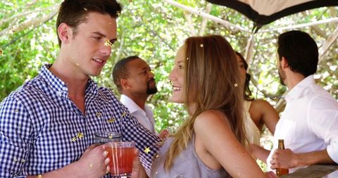Young adults mingling with cocktails under pergola during sunlit backyard gathering