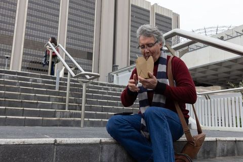 Senior man sitting on urban steps eating takeout with chopsticks, casual commuter lifestyle