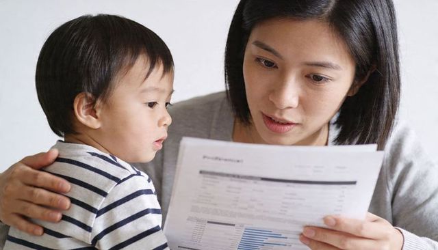 Mother reading household statements while holding toddler, reviewing family finances