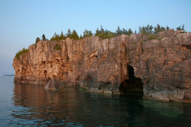 Image shows a majestic rocky cliff with natural arches by the sea, set against a serene backdrop of a forest. The sunset subtly lights the scene, casting a warm glow on the rugged cliffs. Ideal for use in nature tourism promotions, travel blogging, or environmental conservation campaigns.