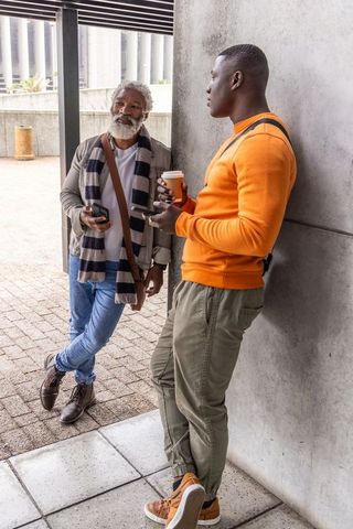 Urban african american men chatting over coffee and smartphones leaning on concrete wall