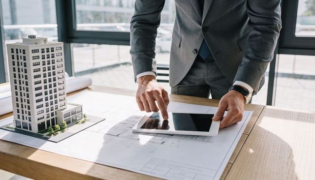 Architect reviewing tablet while pointing at blueprints with building model on desk