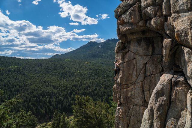 Person engaged in rock climbing on a steep, rugged cliff with expansive green forest and mountain range in background under a bright blue sky with fluffy clouds. The image can be used for promoting adventure sports, outdoor activities, fitness goals, and nature conservation.