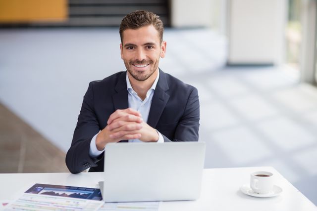 Businessman sitting at desk with laptop and coffee, smiling confidently. Ideal for corporate presentations, business websites, promotional materials, and articles on professional success and productivity.