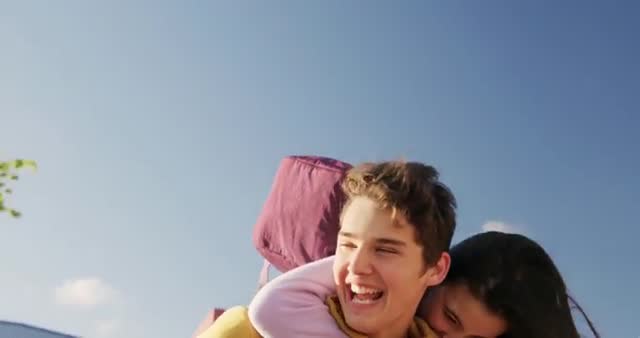 A teenage girl joyfully rides on her boyfriend’s back in front of their school building under a clear sky. The moment is filled with happiness and spontaneity, capturing the carefree joy of young love. Ideal for marketing content aimed at teens, school promotional material, or any media focusing on positive teenage relationships.