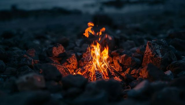 Flickering campfire on rocky shoreline at night casting warm glow and sparks