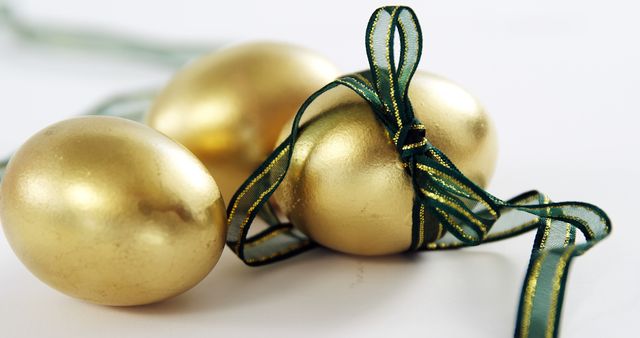 Three gold eggs with a green ribbon on a white background. Ideal for use in Easter promotions, luxury products marketing, holiday decor, and festive celebration themes. The close-up composition highlights the elegance and festive ambiance. Great for cards, advertisements, and seasonal decor blogs.