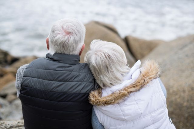 Senior couple sitting on rocks by the beach, enjoying a peaceful moment together. Ideal for use in retirement planning, senior lifestyle, and health and wellness content.