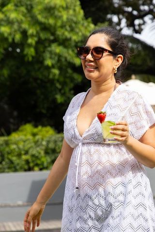 Smiling woman walking poolside in white lace coverup holding mint cocktail with strawberry