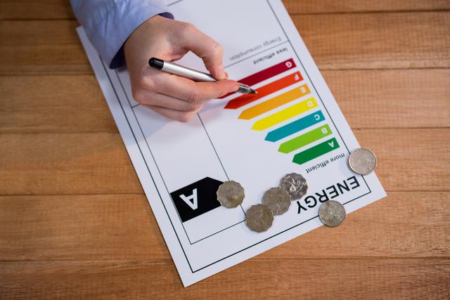 Woman writing on an energy efficiency rating chart with coins scattered on a wooden table. Useful for illustrating concepts related to energy conservation, financial planning, home improvement, and sustainability. Ideal for articles, blogs, and educational materials on energy efficiency and eco-friendly practices.