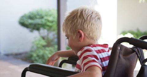 Blond child sitting in wheelchair leaning on porch railing reaching outdoors in daylight
