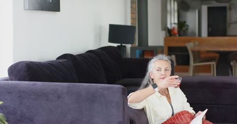 Senior woman relaxing on sectional sofa sipping tea while using tablet in cozy living room