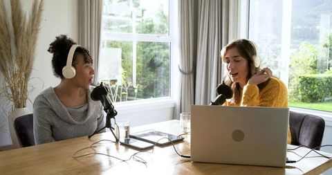 Mid-adult female podcasters recording lively conversation in home studio with laptop