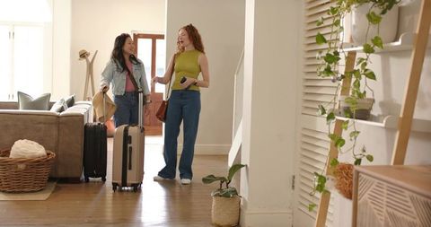 Friends arriving home with suitcases, chatting and checking smartphone in bright living room