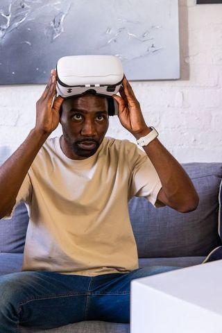 African american man adjusting white vr headset while sitting on gray couch in living room