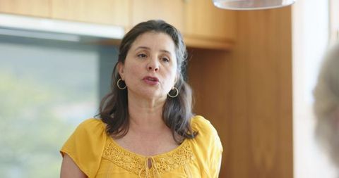 Mature woman speaking in cozy kitchen wearing yellow blouse and gold hoop earrings