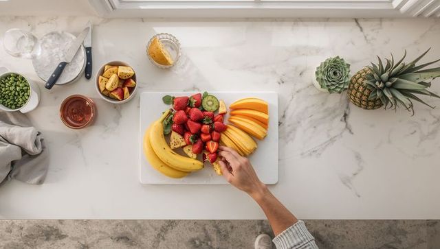 Overhead hand reaching to pick fresh fruit from white cutting board on marble countertop