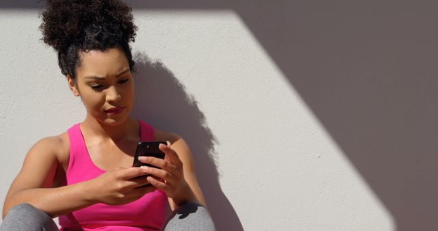 Young woman wearing pink sportswear, sitting indoors in diffused light, focused on using smartphone. Ideal for content related to technology, healthy lifestyle, communication, modern living, and fitness inspiration.