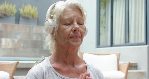 Senior woman meditating with wireless headphones on outdoor patio, practicing mindfulness