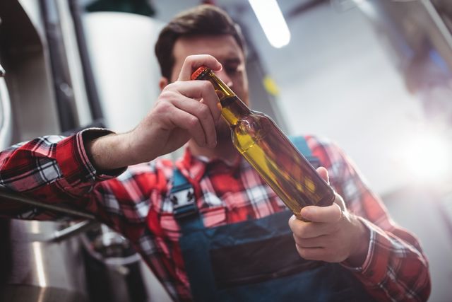 Young brewer in plaid shirt and apron examining beer bottle in brewery. Ideal for content related to craft beer production, quality control in beverage industry, and brewery operations.