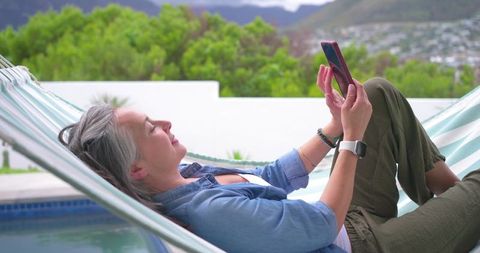 Gray-haired woman relaxing in hammock by pool using smartphone for messaging