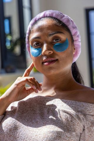 Sunlit indian woman wearing hydrogel under-eye patches, terry headband, at-home skincare routine