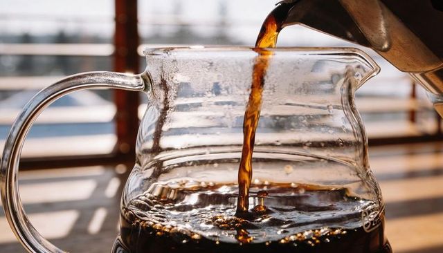 Pouring dark coffee into clear glass carafe with sunlit ripples on wooden counter