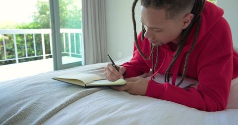 African american man journaling on bed in red hoodie, sunlit bedroom balcony and greenery