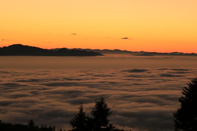 Breathtaking view of the sunset over a mountain range with a sea of clouds beneath the peaks. The sky has a beautiful orange hue, creating a tranquil and serene atmosphere. This image is ideal for use in travel brochures, nature websites, and desktop wallpapers.