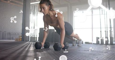 Athletic woman holding plank gripping hex dumbbells in industrial backlit gym for strength training