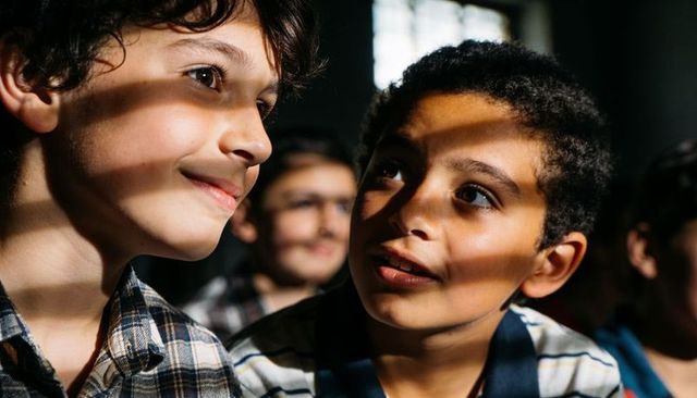 Two schoolboys sharing quiet moment with striped window light and thoughtful expressions