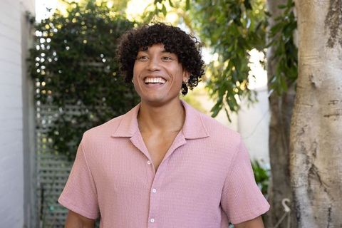 Smiling young man with curly hair standing in sunlit courtyard wearing pink collared shirt