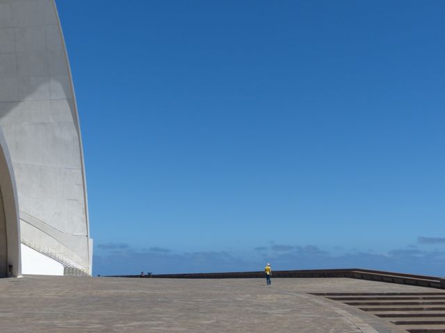 Stunning shot of modern architectural structure with clean lines and concrete surfaces against a backdrop of clear blue sky. A solitary person wearing a yellow hard hat emphasizes scale and minimalistic design. Perfect for use in themes around architecture, construction, tourism, and modern design.