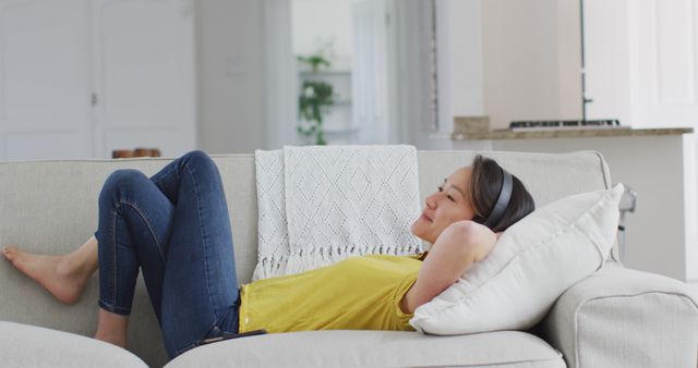 Smiling young woman lying on beige couch in living room. Wearing yellow shirt and jeans, she appears comfortable and at ease. Perfect for themes of relaxation, home life, well-being, casual lifestyle, and solitude. Suitable for use in home decor promotions, lifestyle blogs, or advertisements for comfortable clothing or home products.