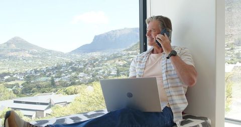 Mature man working remotely on laptop talking on phone at window seat with scenic valley view
