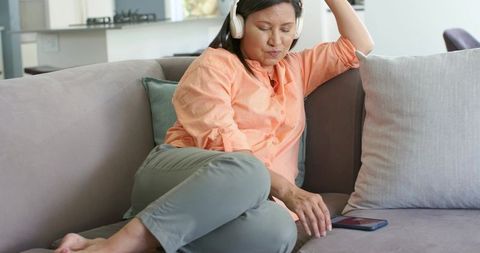 Mature asian woman reclining on gray sofa wearing headphones reaching for smartphone