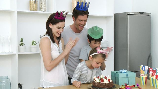 Family celebrating daughter's birthday in kitchen. Little girl blowing out candles on cake while parents and brother smile. Perfect for family life, celebrations, birthdays, and home gatherings themes.