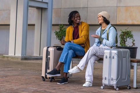 Young diverse travelers smiling and chatting on bench with luggage in urban plaza