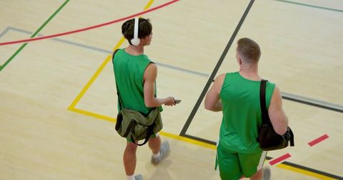 Two male teammates walking gym court carrying bags and gearing up for basketball training