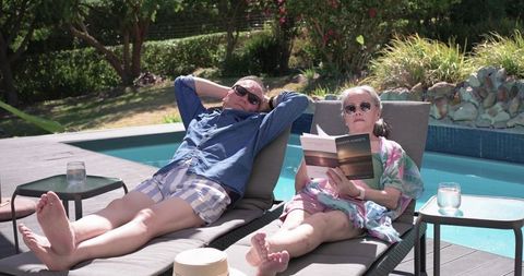 Asian mature couple relaxing poolside on lounge chairs reading paperback and sunbathing