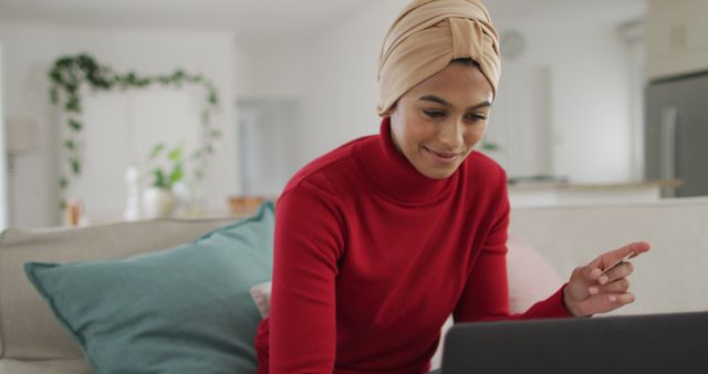 Image of happy biracial woman in hijab sitting on sofa and using laptop. Lifestyle, spending free time at home with technology concept.