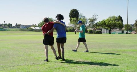 Diverse teammates jogging, celebrating after football drill in sunny park showing teamwork